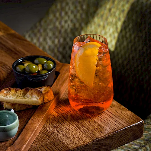 A wooden board holds an orange iced cocktail with ice, olives in a bowl, bread, and a small green cup beside it.
