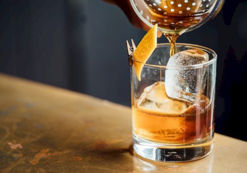 A glass of whiskey with large ice cubes, a cherry-orange twist, and a cocktail being poured into it from a shaker, on a wooden bar.