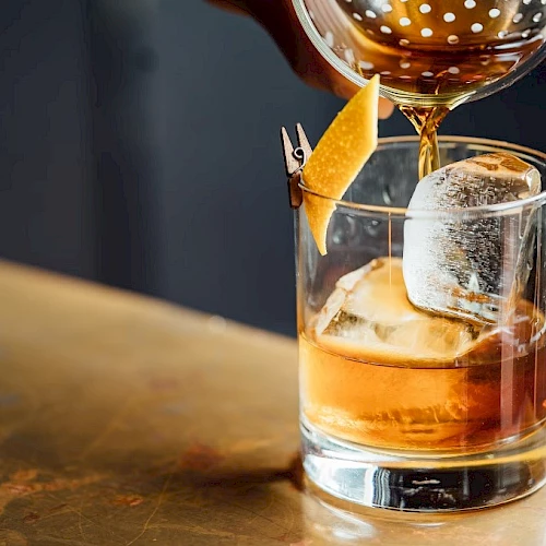 A glass of whiskey with large ice cubes, a cherry-orange twist, and a cocktail being poured into it from a shaker, on a wooden bar.