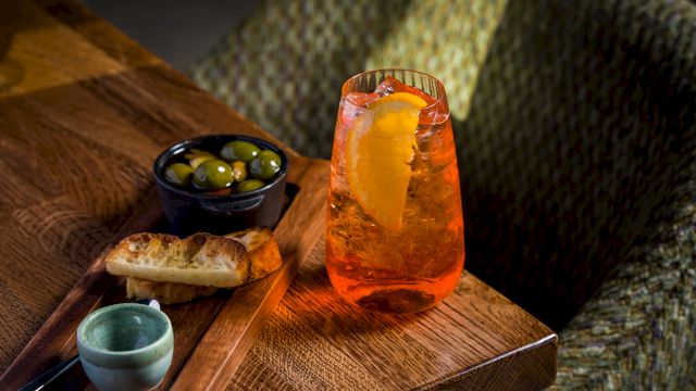 A wooden board holds an orange iced cocktail with ice, olives in a bowl, bread, and a small green cup beside it.