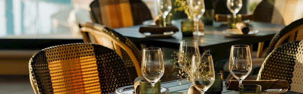 A stylish outdoor dining setup with a dark table, wicker chairs, wine glasses, candles, and a sunny poolside background.