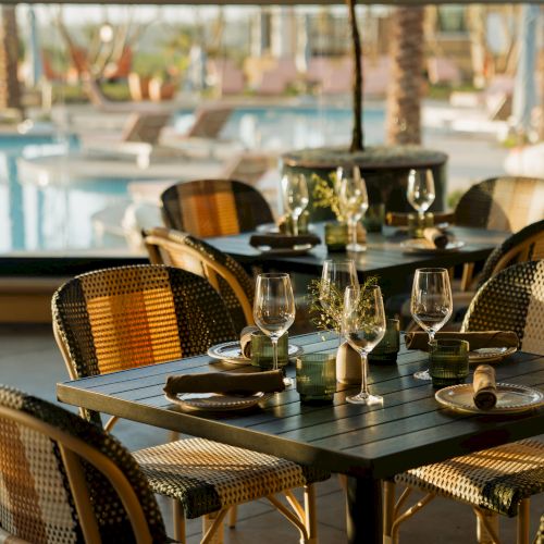 A stylish outdoor dining setup with a dark table, wicker chairs, wine glasses, candles, and a sunny poolside background.