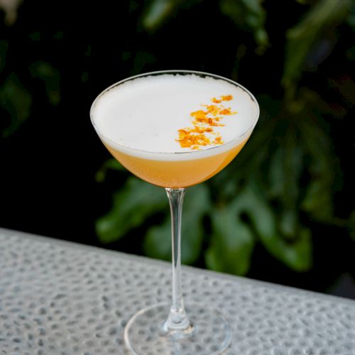 A white cocktail glass with an orange garnish on a long stem, placed on a dotted surface outdoors.