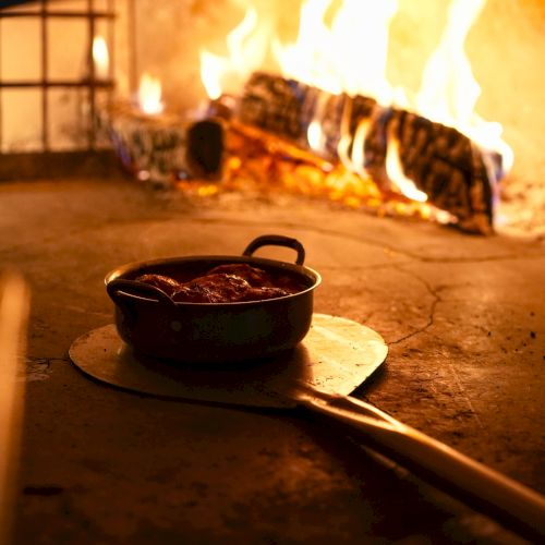A small pot of food sits on a stone table as a large fire or grill burns in the background.