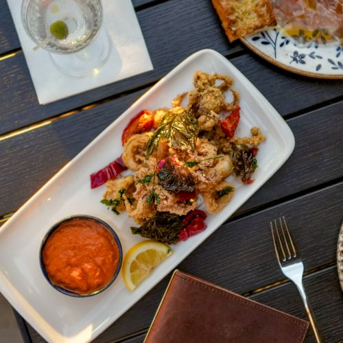 A plated meal with fried chicken or seafood, roasted peppers and greens, dipping sauce, lemon, bread, and a drink on a restaurant table.