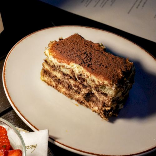A slice of chocolate tiramisu cake with cocoa dusting sits on a white plate.