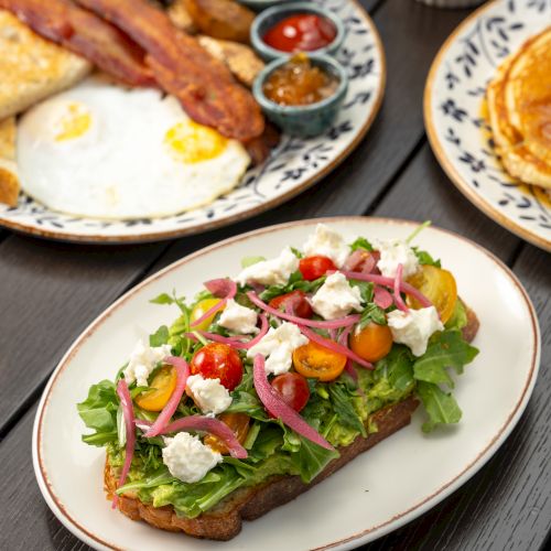 Plates of breakfast and salad toast&mdash;sunny-side eggs, sausages with dips, and a greens-and-cheese toast topped with tomatoes and onions.