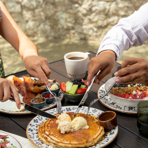 An outdoor breakfast table with pancakes, fruit bowls, coffee, and people using forks beside a river.