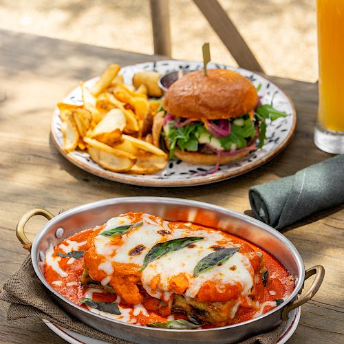 A table meal: a loaded burger, golden fries, a skillet of cheesy tomato casserole, and a glass of orange drink.