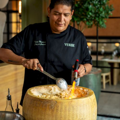 A chef in a dark &ldquo;VERISE&rdquo; shirt serves food with a spoon from a big cheese wheel outdoors at night.