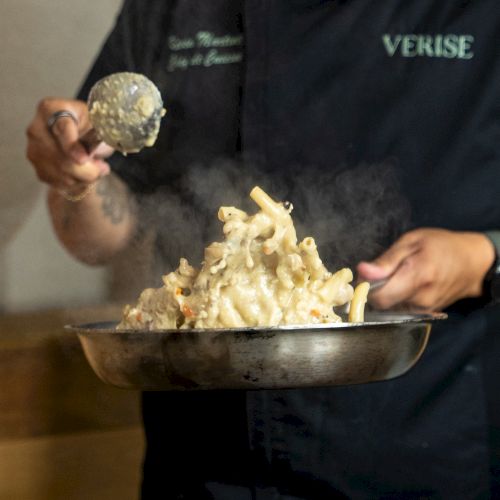 A person in a &ldquo;VERISE&rdquo; hoodie lifts cheesy pasta from a metal bowl, ready to serve, with foil in hand.