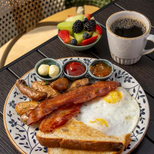 A hearty breakfast plate with bacon, sausages, fried eggs and toast, plus a fruit bowl, three sauces, and a cup of coffee.