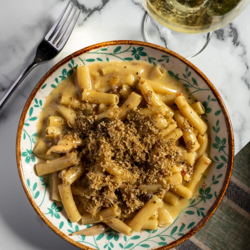 A bowl of creamy penne pasta topped with grated cheese sits beside a fork and a glass of white wine.