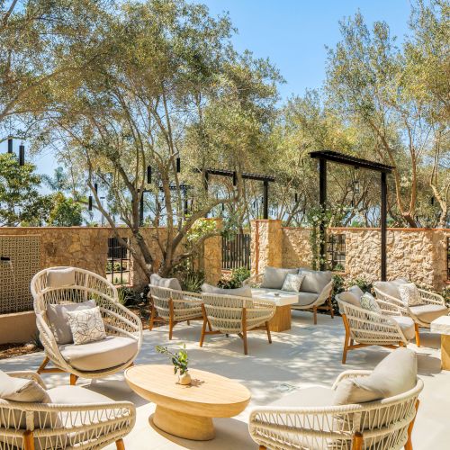 A sunny outdoor patio with wicker lounge chairs and tables, framed by trees, stone walls, and shaded pergolas.
