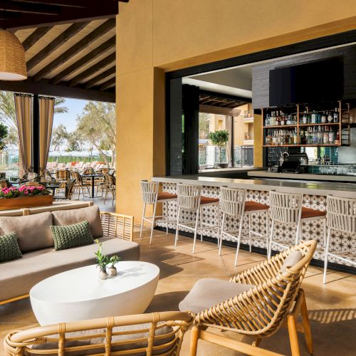 An elegant outdoor terrace with rattan lounge seating, a stocked bar counter, and palm-lined views.