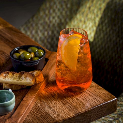 A wooden board holds an orange iced drink with citrus slices, a bowl of green olives, and bread/rolls beside it.