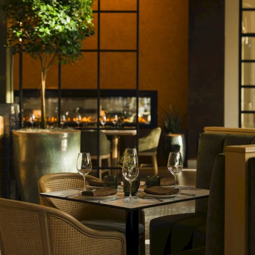 A warmly lit restaurant patio table set with wine glasses, cutlery, candles, and green plants beside a large window.
