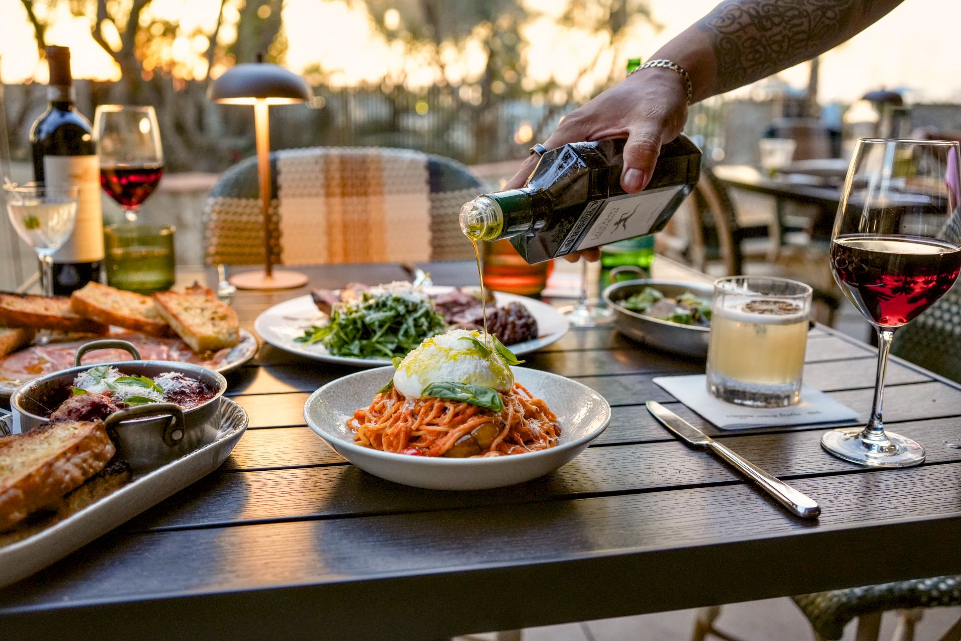 At an outdoor dinner, a hand pours wine while plates of salad, bread, and sauces sit on a patio table.