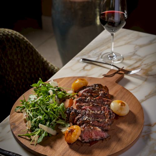 A plate with two medium-rare steaks, topped with greens, a side of mashed potatoes, and a glass of red wine on a rustic wooden table.