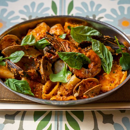 A pot of pasta with tomato sauce, basil leaves, and mushrooms served in a metal pan on a white tray, colorful patterned table beneath.