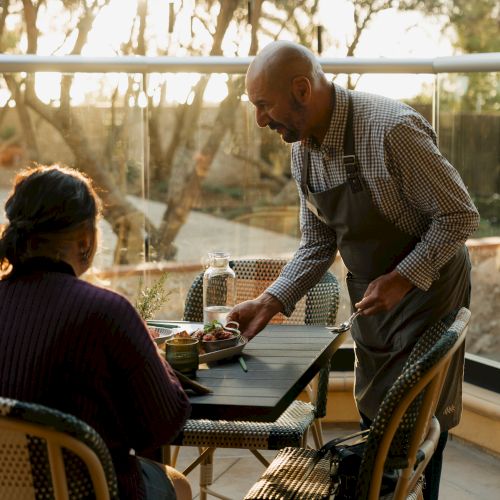 A man in an apron serves food to a seated woman on a balcony at sunset, with trees and warm light in the background.