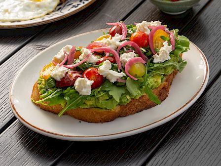 Open-faced salad toast with greens, tomatoes, onions, and goat cheese, alongside fruit cups and sausages with dipping sauces.