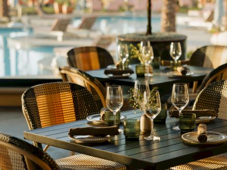 A stylish outdoor restaurant setup with a square table set for four, wine glasses, plates, and cutlery, poolside backdrop, warm afternoon light ending the sentence.