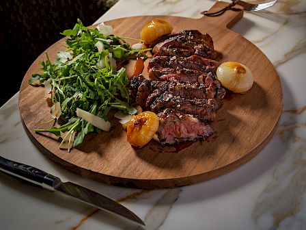 A sizzling steak with greens and roasted garlic on a round board, paired with a glass of red wine and a marble table top.