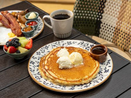 Breakfast spread: fluffy pancakes with butter, bacon and eggs, fruit bowl, sauces, and two cups of coffee for a cozy morning.