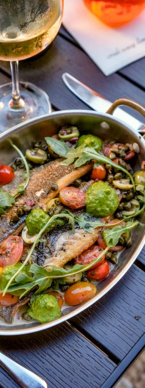 A metal pan of grilled fish with greens, cherry tomatoes and herbs, served at a table beside a wine glass.