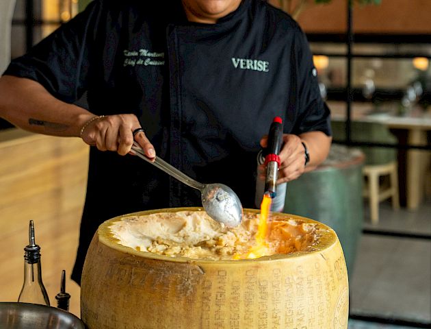 A chef in a dark uniform serves food from a large cheese wheel, using a spoon, with utensils on a table.