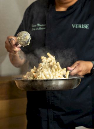 A person in a dark &ldquo;VERISE&rdquo; hoodie eats cheesy pasta from a metal bowl, lifting it with a fork like garlic bread.