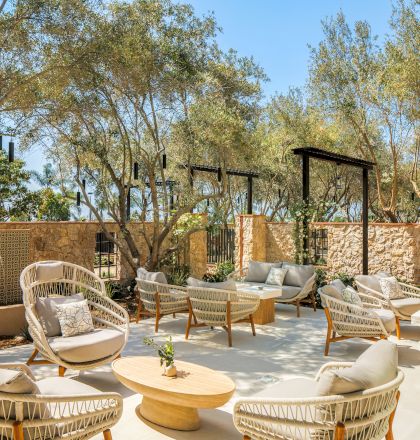 A sunny outdoor patio with wicker lounge chairs and tables, stone walls, lush trees, and modern garden structures under a blue sky.