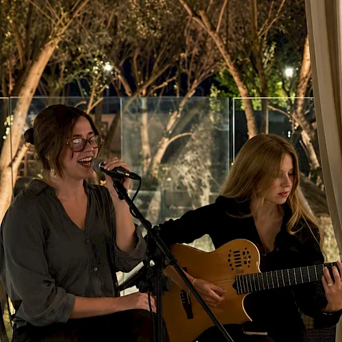 A woman sings into a microphone while another plays an acoustic guitar at an outdoor evening performance.