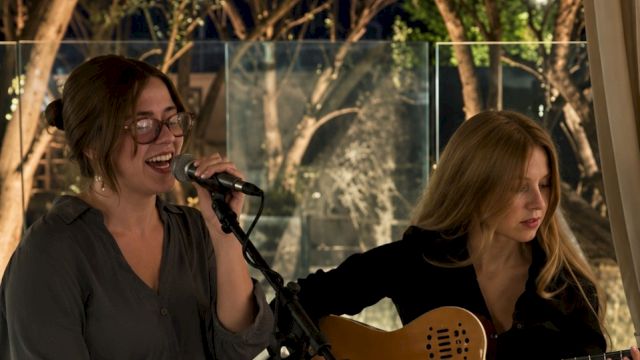 A woman sings into a microphone while another plays an acoustic guitar at an outdoor evening performance.